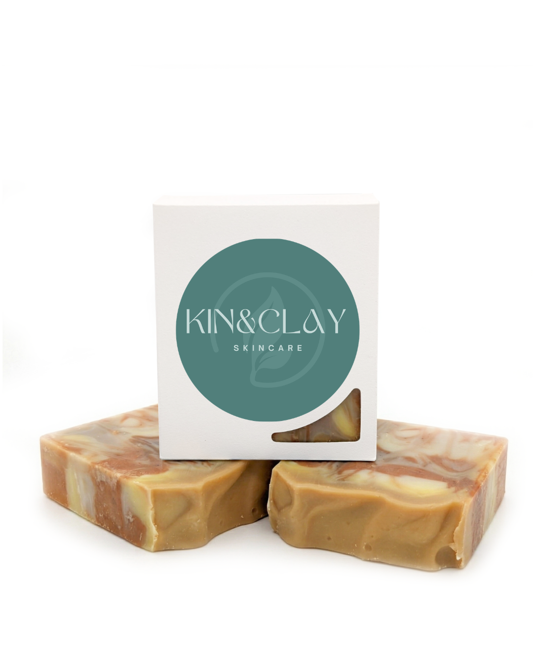 Two tan swirled bars of Kin & Clay Sandalwood Clay Soap stacked in front of their branded box